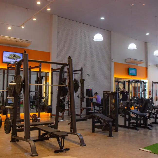 Academia moderna de personal training com equipamentos de musculação, racks e bancos, pessoas treinando, ambiente otimizado para resultados.