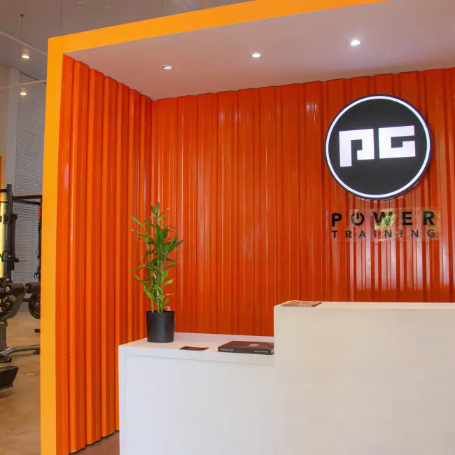 Recepção moderna de estúdio Power Training com balcão branco, planta e logo PG em parede laranja, indicando personalização.