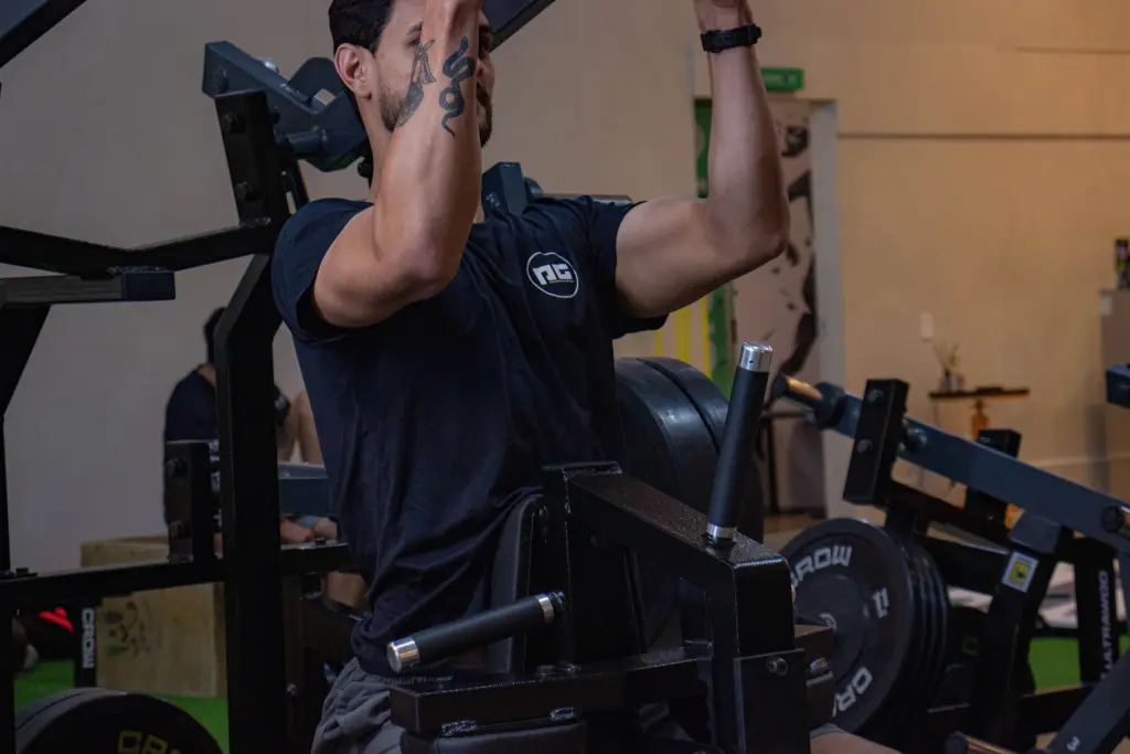 Homem com tatuagens treinando força em máquina de musculação na academia, focado em personal training e resultados eficazes.