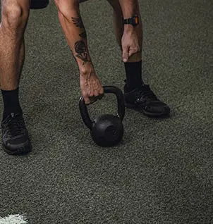 Homem segurando kettlebell preto no chão do estúdio de personal training. Treino funcional e condicionamento físico personalizado.